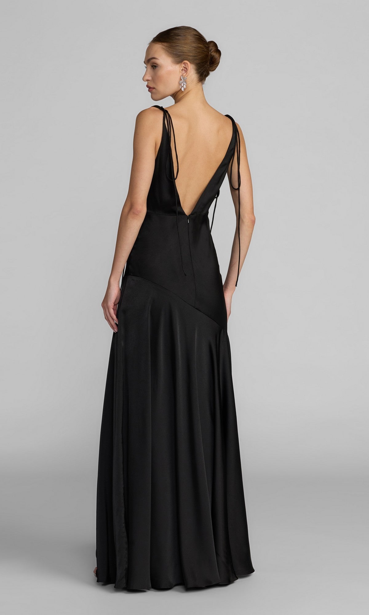 A woman stands facing away from the camera against a plain background, wearing the Mac Duggal 49914 long formal dress—a black, floor-length satin gown featuring an elegant A-line silhouette, deep V-back, and tied shoulder straps.